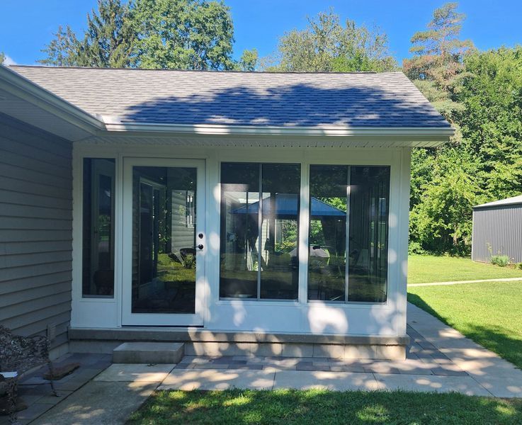 Sunroom addition with white trim, glass windows, and a gray roof, attached to a house with beige siding.