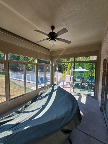 Covered table in a screened porch with a ceiling fan, windows overlooking a yard and pool.