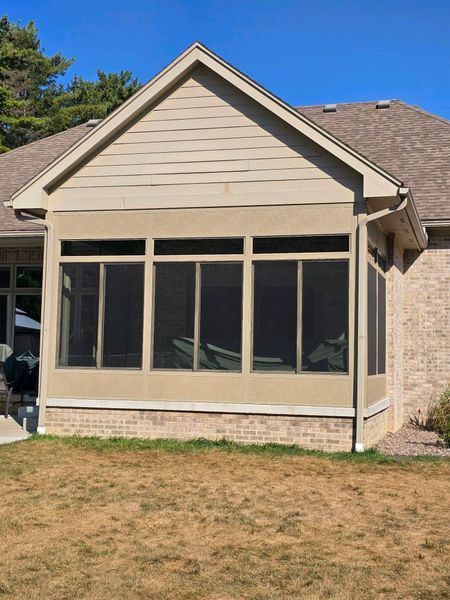 Screened porch with tan siding, brick base, and brown roof, attached to a brick home on a sunny day.