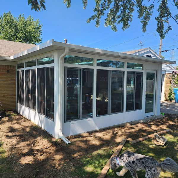 White sunroom addition with large windows and a screen door, attached to a brick home.