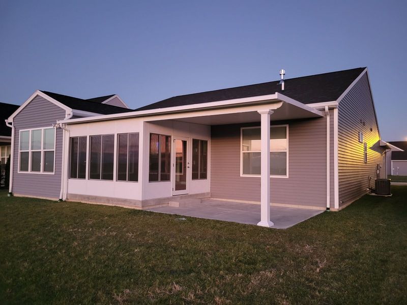 Back of a house with attached screened-in porch and patio, gray siding, green lawn, evening light.