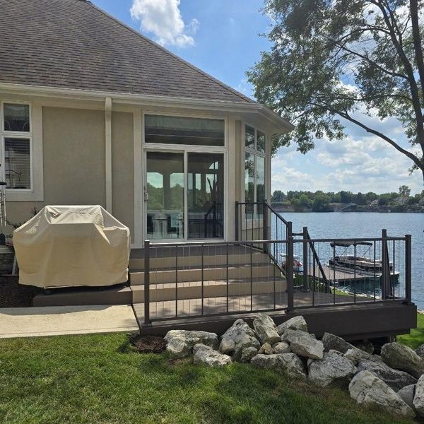 Deck overlooking a lake; beige house with sliding glass door, brown railing, covered grill, and rocks.