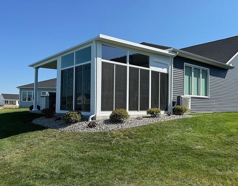 Sunroom addition with screen panels, attached to a house with blue siding, on a grassy hill under a blue sky.