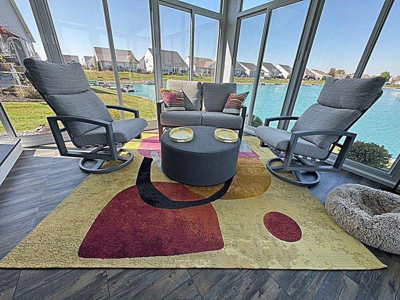 Sunroom with modern furniture: two swivel chairs, sofa, coffee table, colorful rug, lake view.