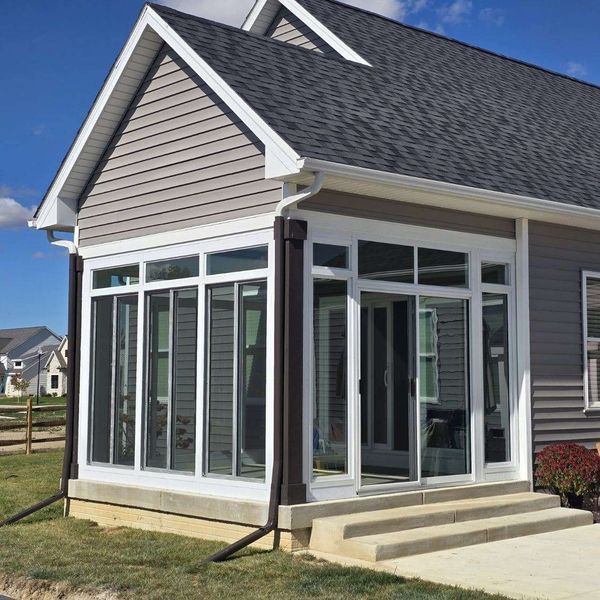 Sunroom with white-framed windows, gray siding, and a dark shingle roof. Steps lead to a concrete patio.