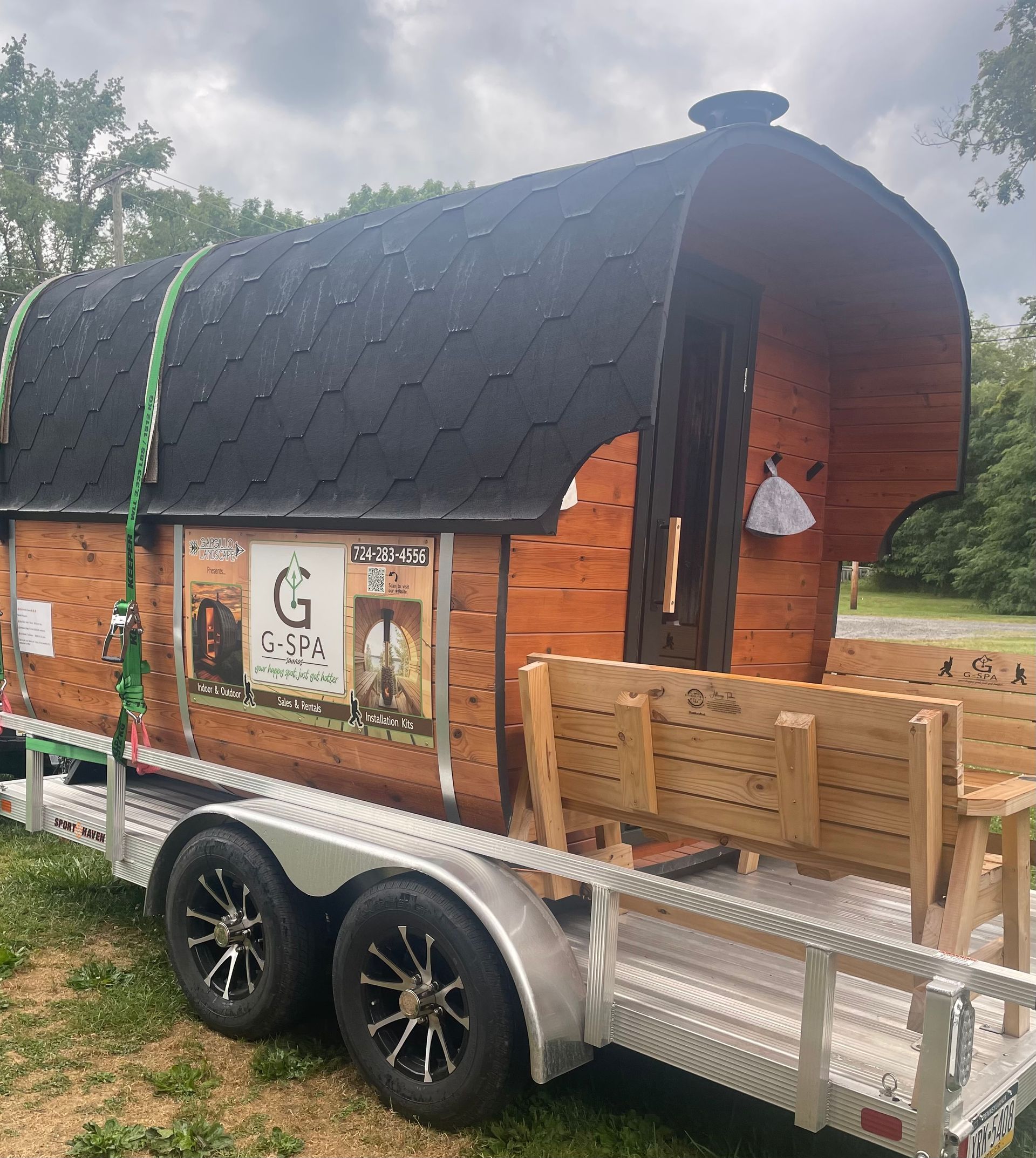 Wooden sauna on a trailer with black roof, benches, and a business advertisement.
