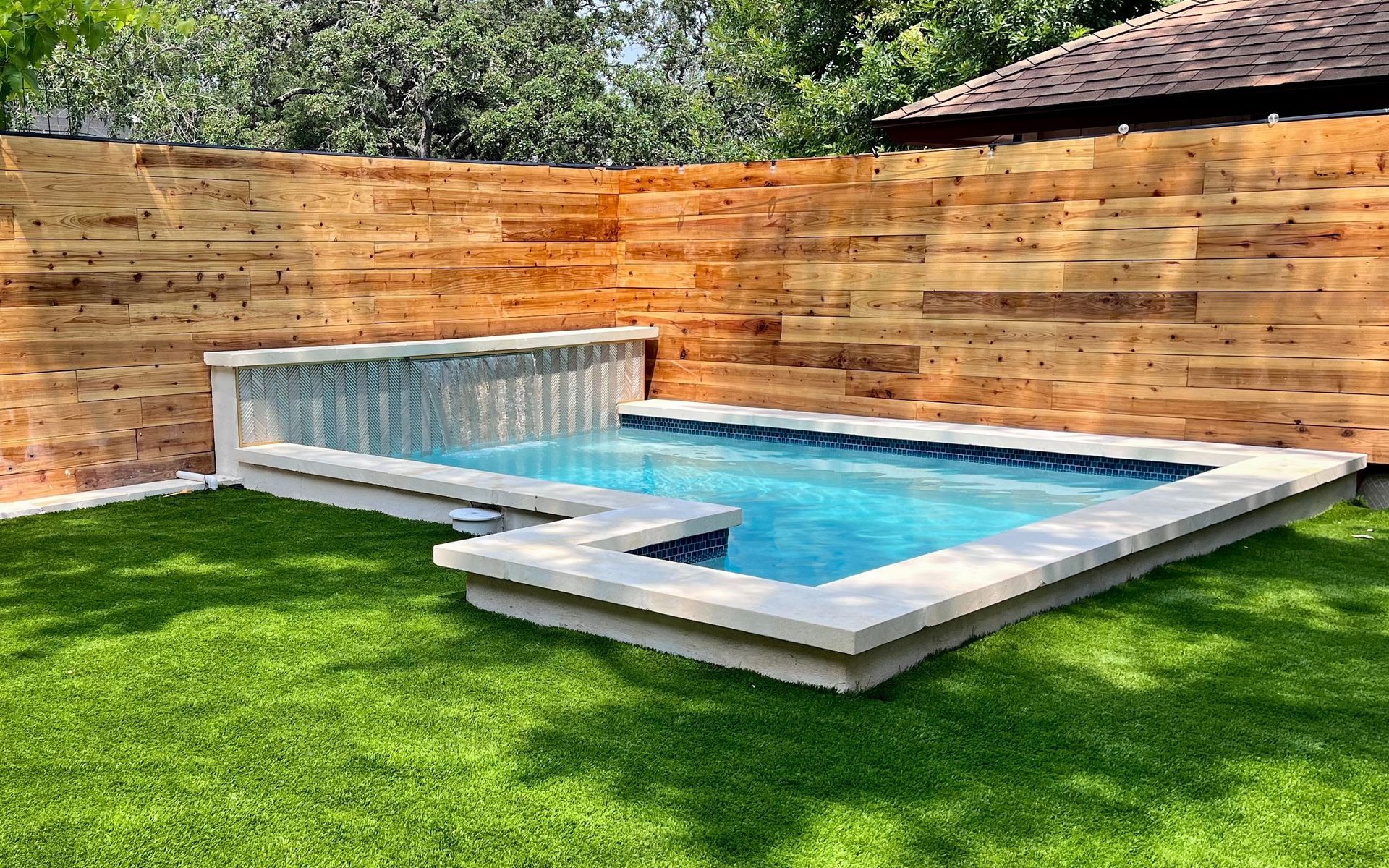 A small pool with a waterfall feature, bordered by a wooden fence and artificial grass.
