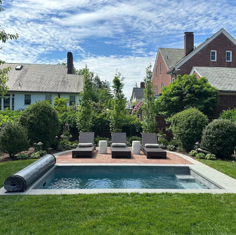 A small pool with lounge chairs, brick patio, and lush landscaping in a backyard setting.