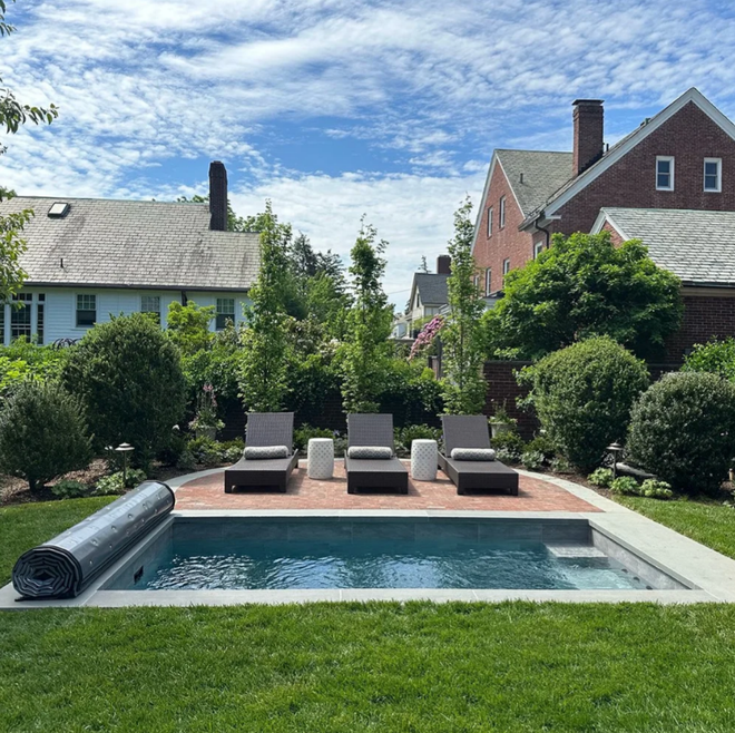 Small rectangular pool with lounge chairs on a brick patio in a backyard with surrounding greenery and houses.