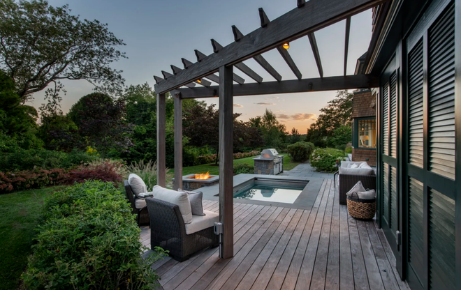 Wooden deck with pergola, seating, fire pit, small pool, and surrounding lush garden at dusk.