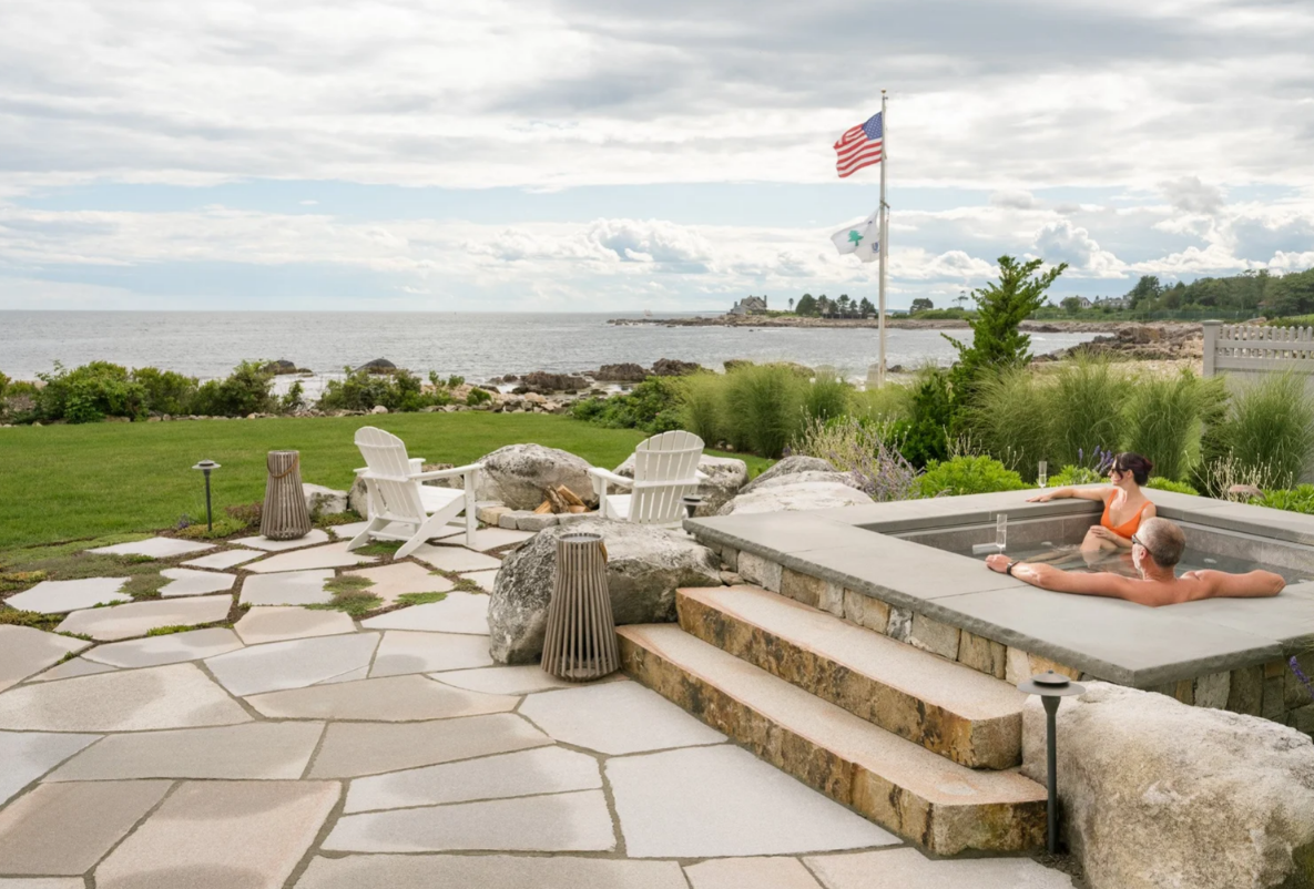 Couple in a hot tub overlooking the ocean, with an American flag and stone patio.