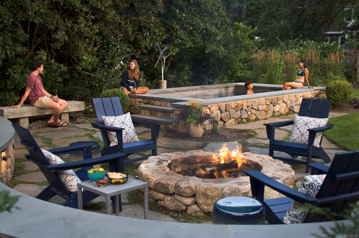 People relaxing around a fire pit and pool in a backyard. Blue chairs, stone patio, lush greenery.