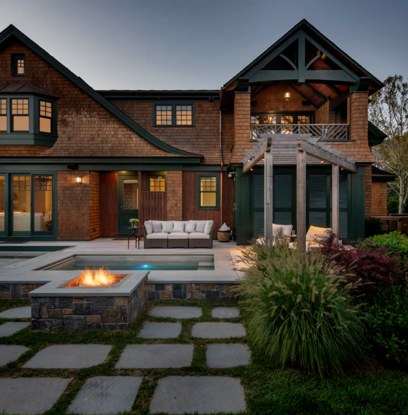 A luxurious home exterior with a pool, fire pit, and patio. Brown shingles, greenery, and a pergola are visible.