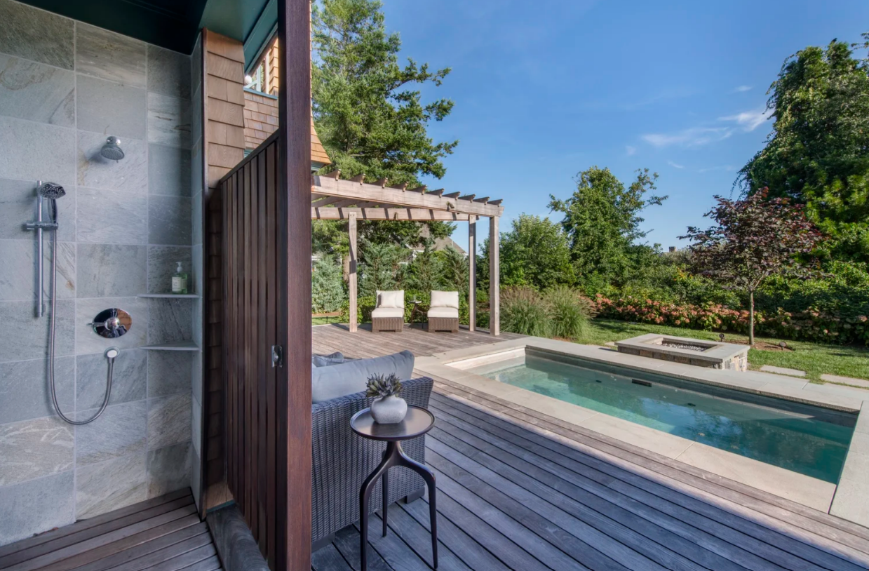 Outdoor shower next to a small pool, deck, and seating area, with trees and sky in the background.