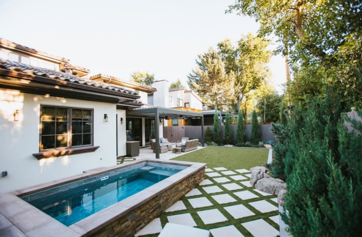 Backyard with pool, tiled patio, green lawn, trees, and a home.