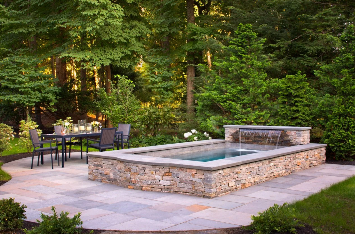 Stone-walled pool with waterfall feature next to a patio with a table and chairs, surrounded by trees.