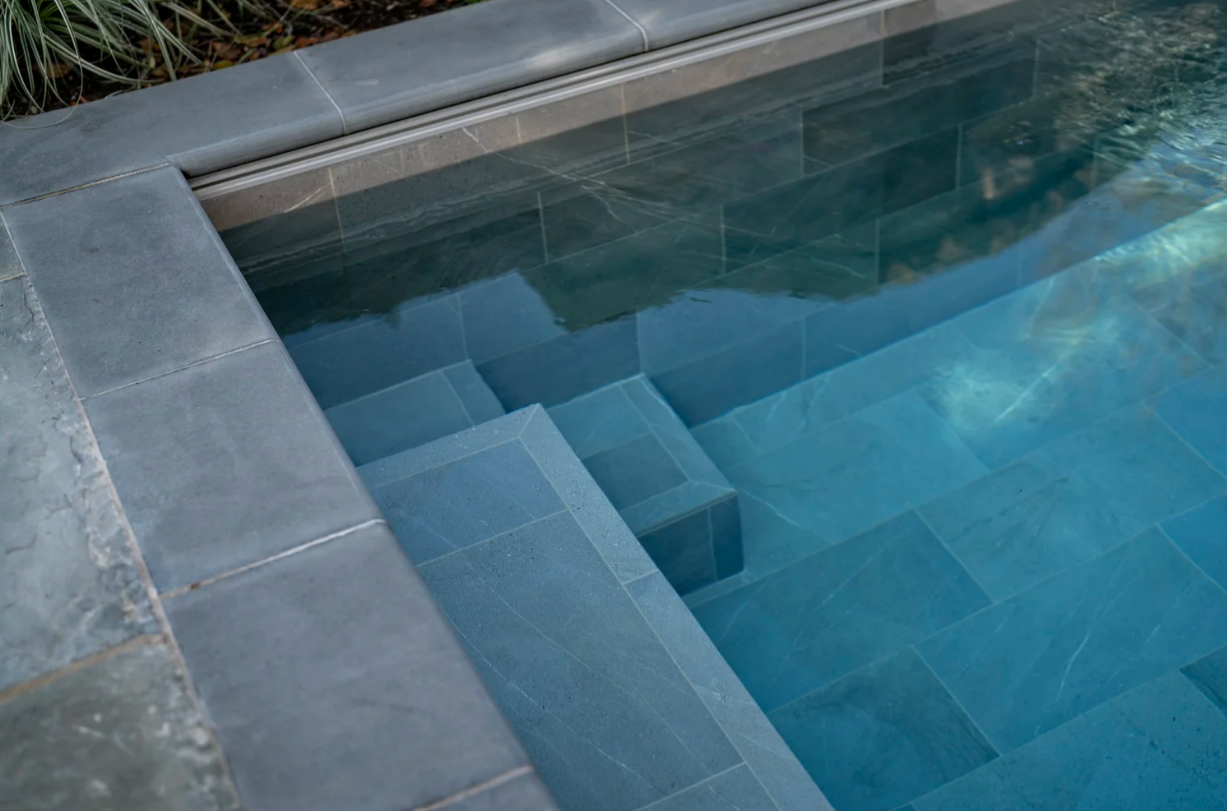 Pool steps with blue-gray tile submerged in clear water; a linear drain is visible at the top.