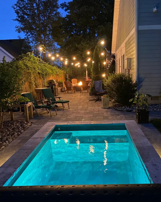 Nighttime backyard scene with illuminated pool, string lights, and fire pit.