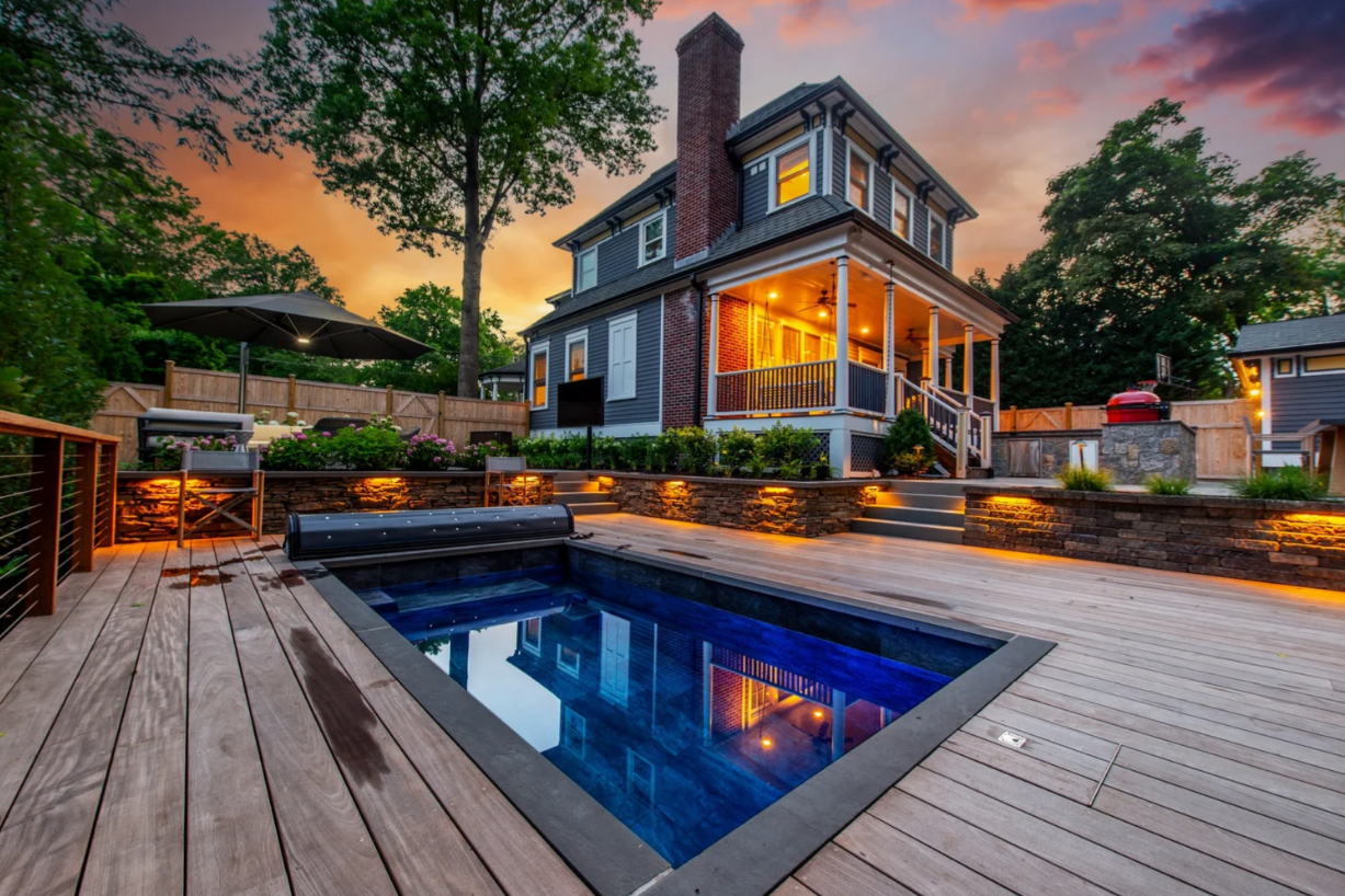 Pool deck with a dark blue pool, next to a grey house with a porch; sunset sky.