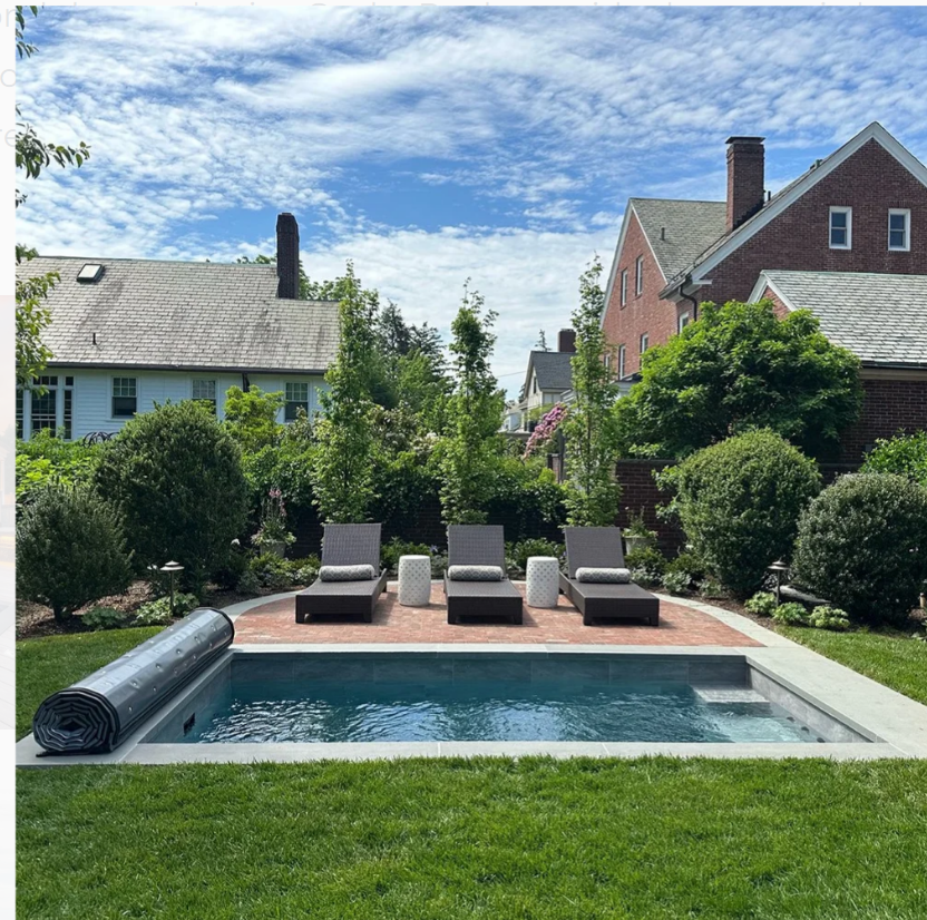 A backyard pool with lounge chairs on a brick patio, surrounded by green grass and trees, with houses in the background.