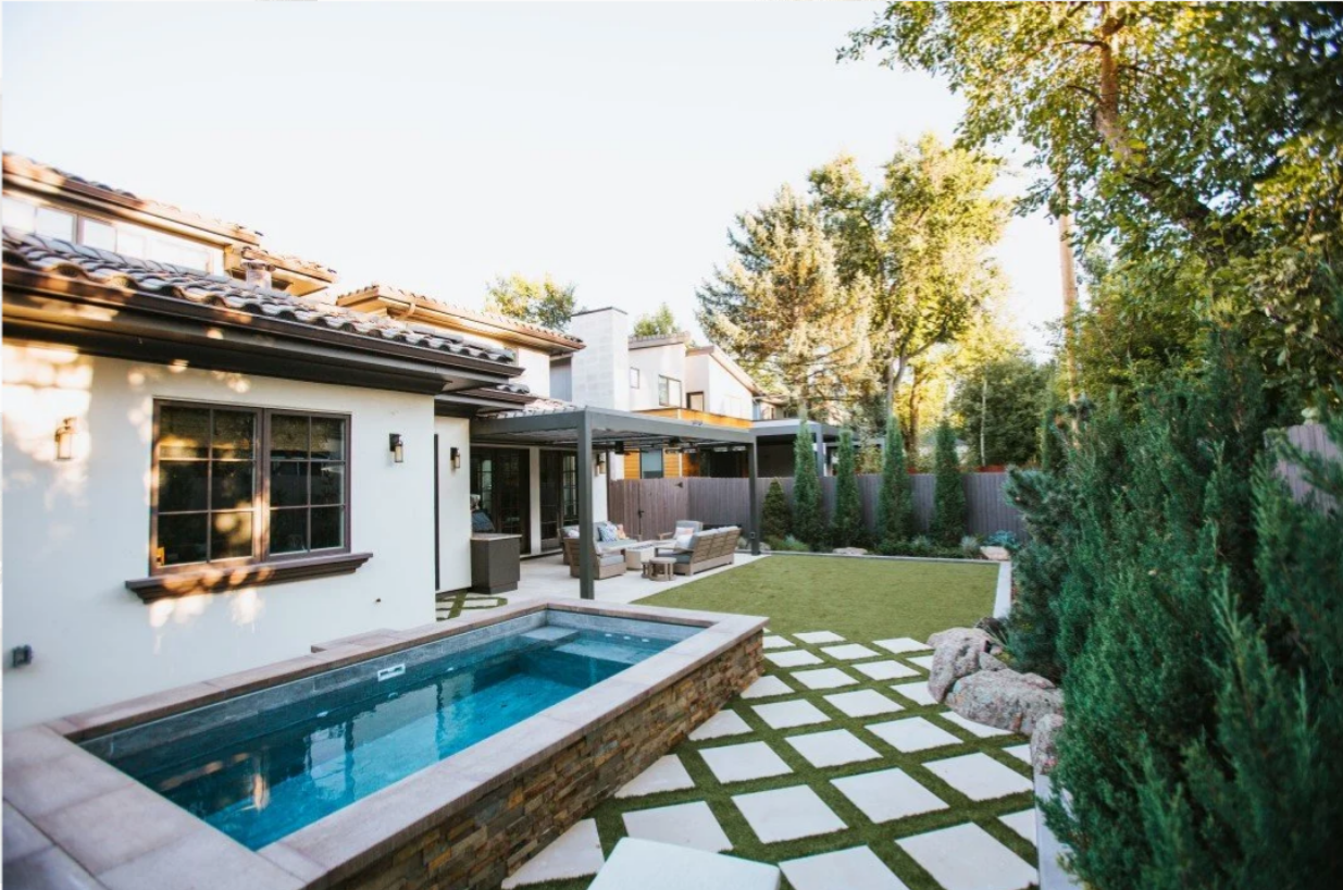 Backyard with rectangular pool, patio, and green lawn; house with light-colored exterior and trees in the background.