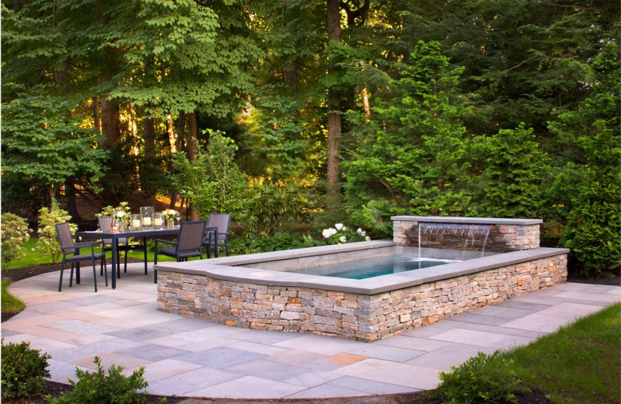 Outdoor patio with rectangular pool, stone wall, waterfall, and dining table. Green trees in background.