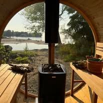 Inside a sauna, a black stove sits beneath a window overlooking a lake.