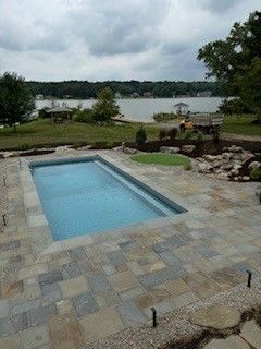 Rectangular pool with blue water and stone patio overlooking a lake on a cloudy day.
