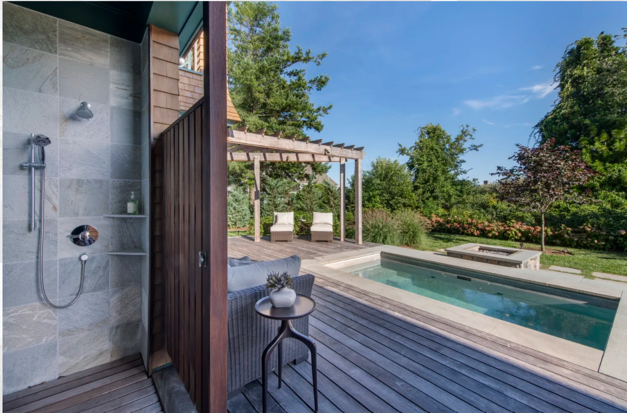 Outdoor shower, small pool, and seating area in a backyard setting with wooden deck and blue sky.