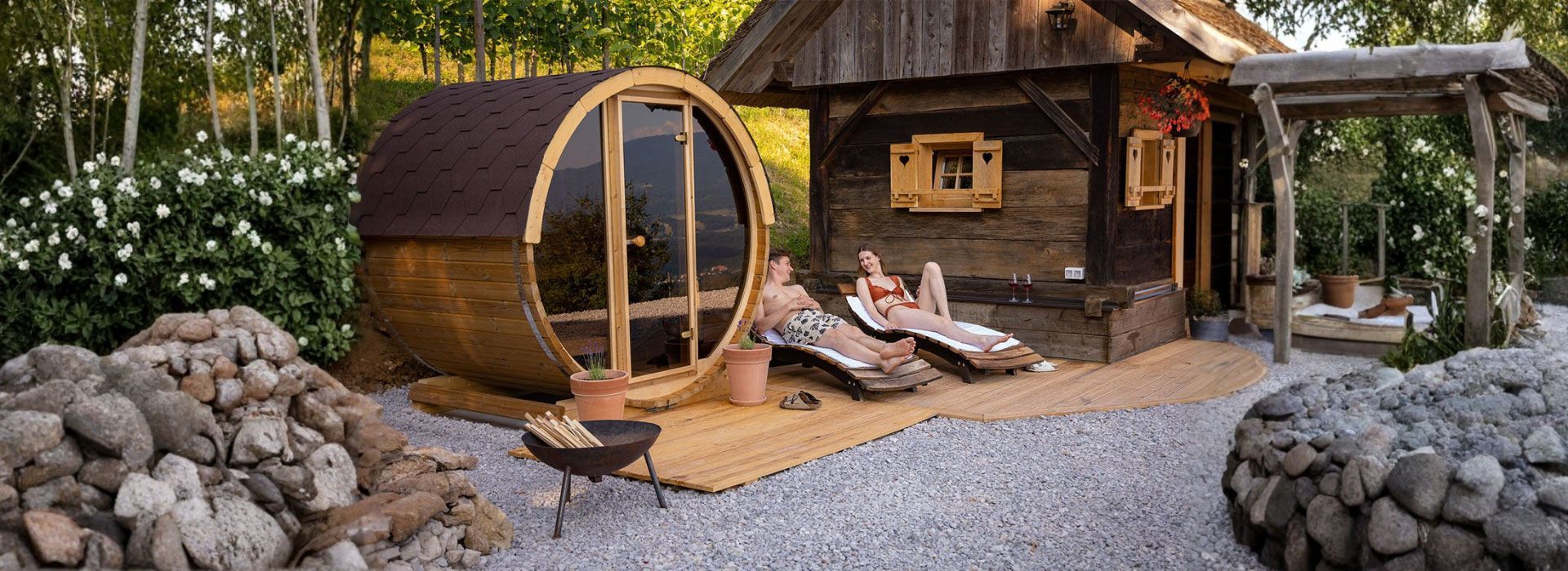 A barrel-shaped sauna and wooden cabin with people lounging on chairs, surrounded by greenery.
