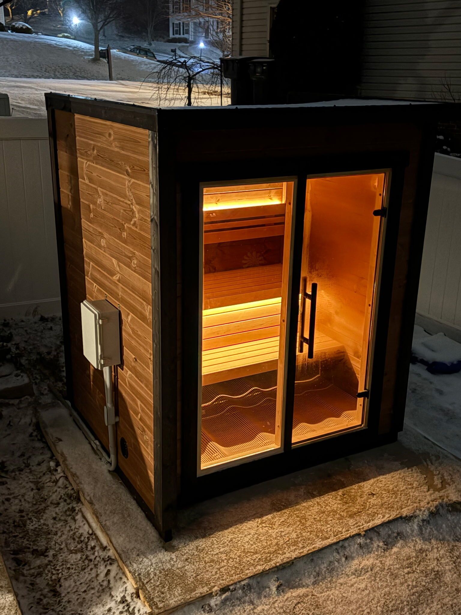 A small outdoor wooden sauna with a glowing, lit glass door sits on a snowy patio at night.