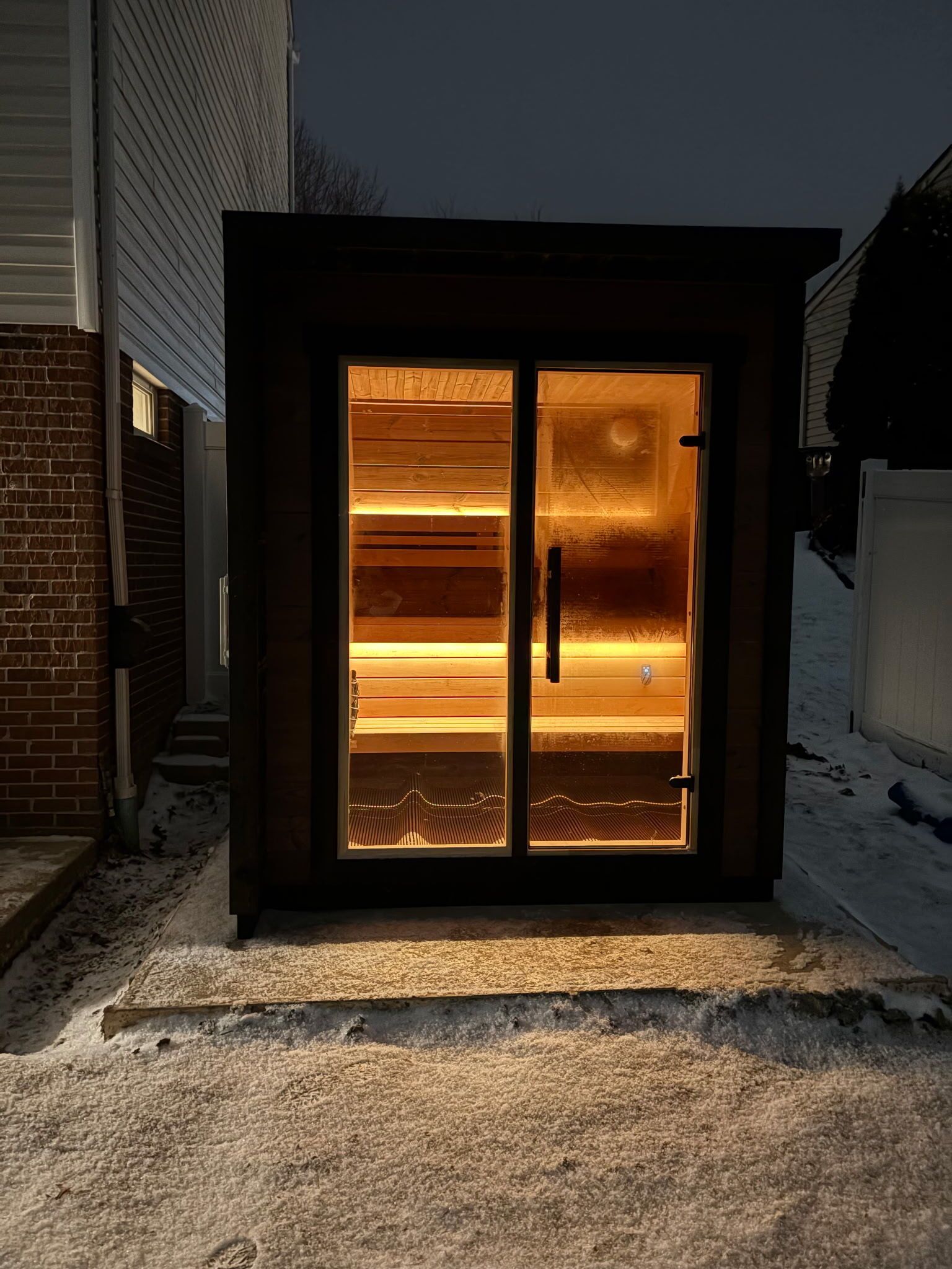 An illuminated outdoor sauna with glass doors, set against a dark, snowy backyard at night.