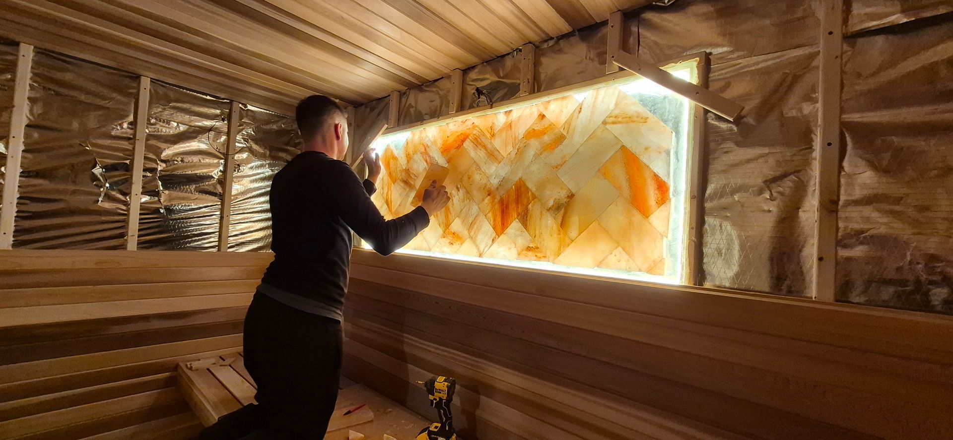 A person kneeling in a wooden sauna, installing a backlit salt brick wall panel.