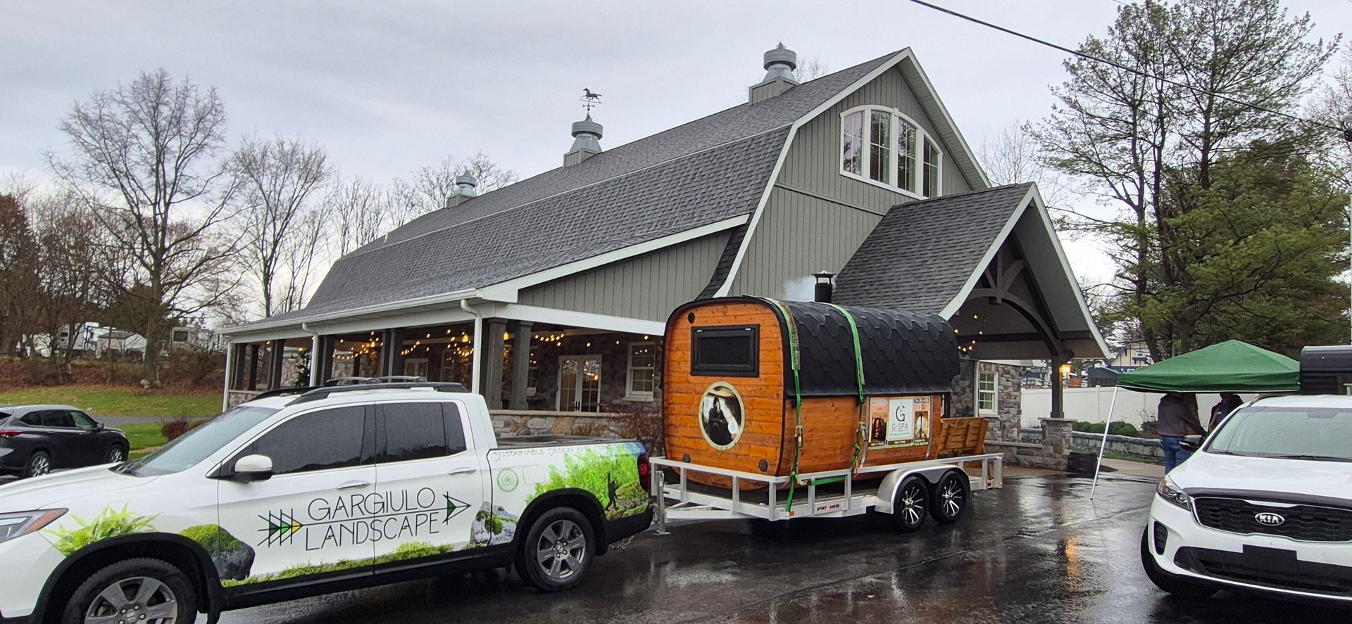 A white pickup truck hauls a wood-paneled sauna trailer in front of a large barn-style building on an overcast day.