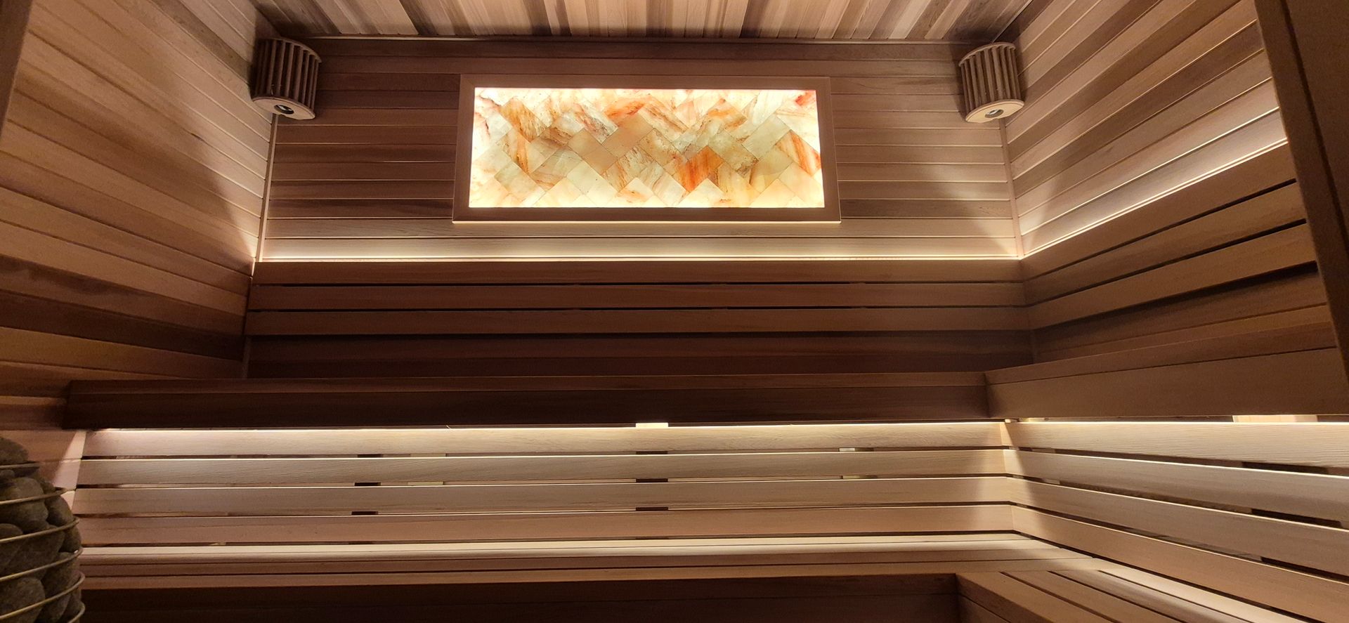 Modern sauna interior with wood-paneled walls, tiered benches, warm accent lighting, and a backlit salt block panel.