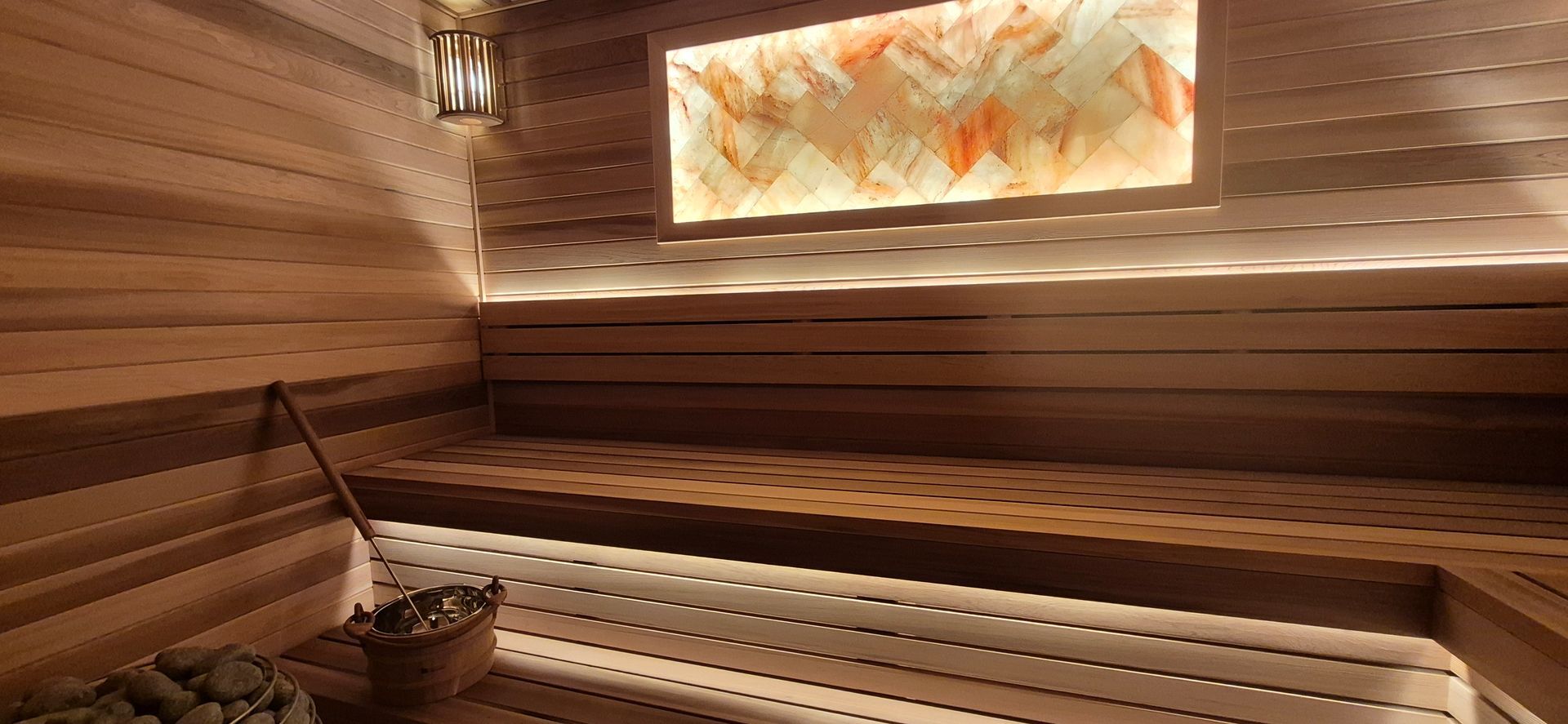 A warm-toned wooden sauna featuring slatted benches, a stone heater, and an illuminated Himalayan salt brick wall panel.