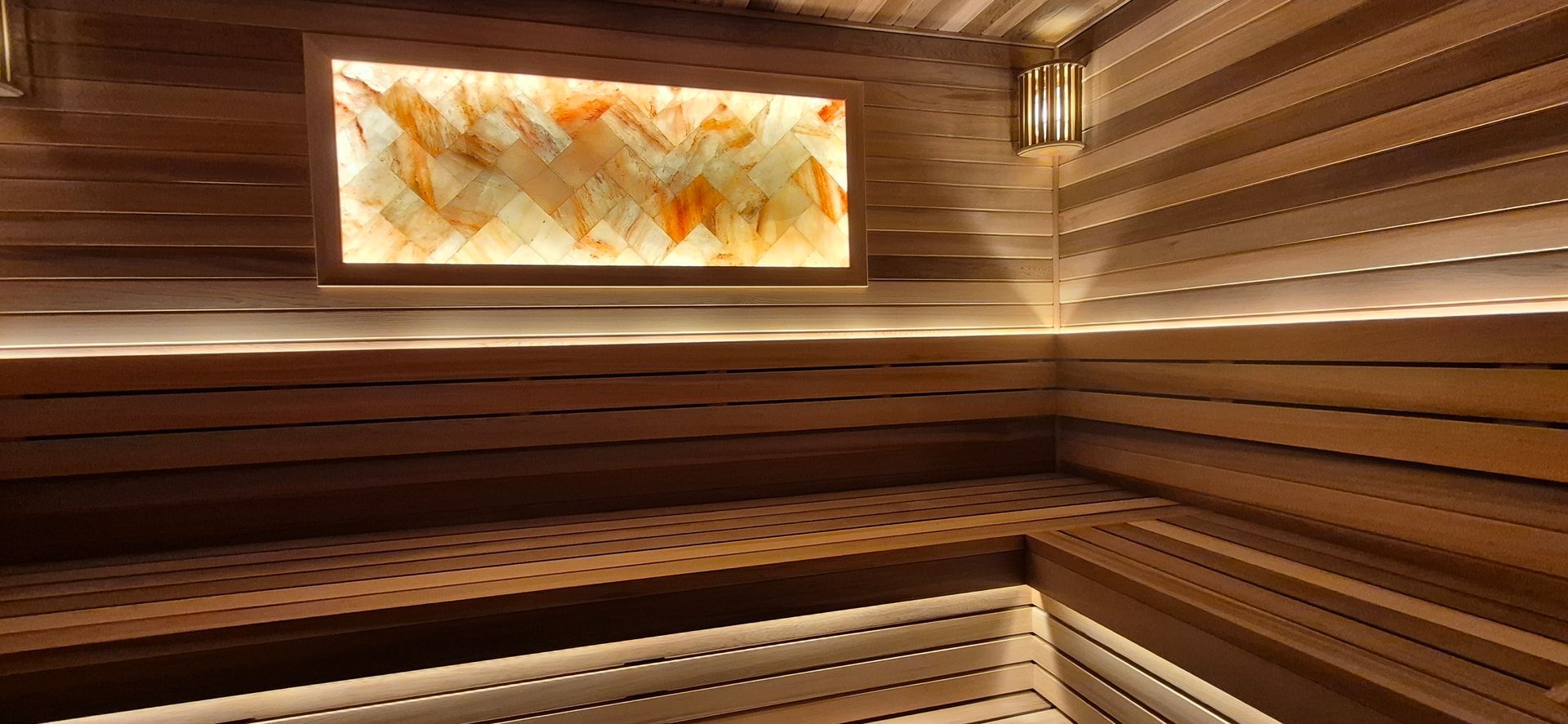 Interior view of a modern wooden sauna featuring tiered bench seating and an illuminated salt brick accent wall.