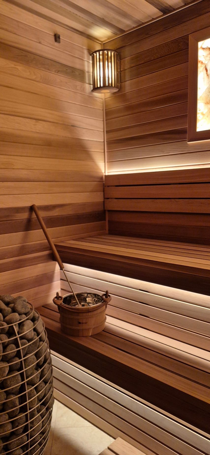 A modern sauna interior with tiered wooden benches, warm accent lighting, a wooden bucket with a ladle, and a stone heater.
