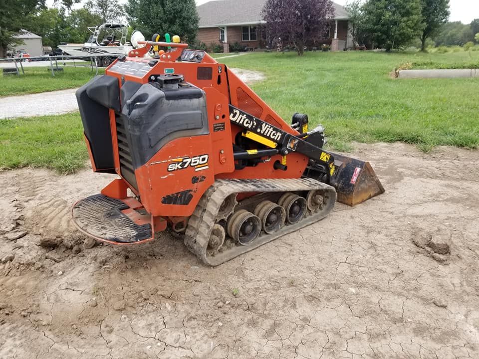 A small orange and black machine is sitting in the dirt in front of a house.