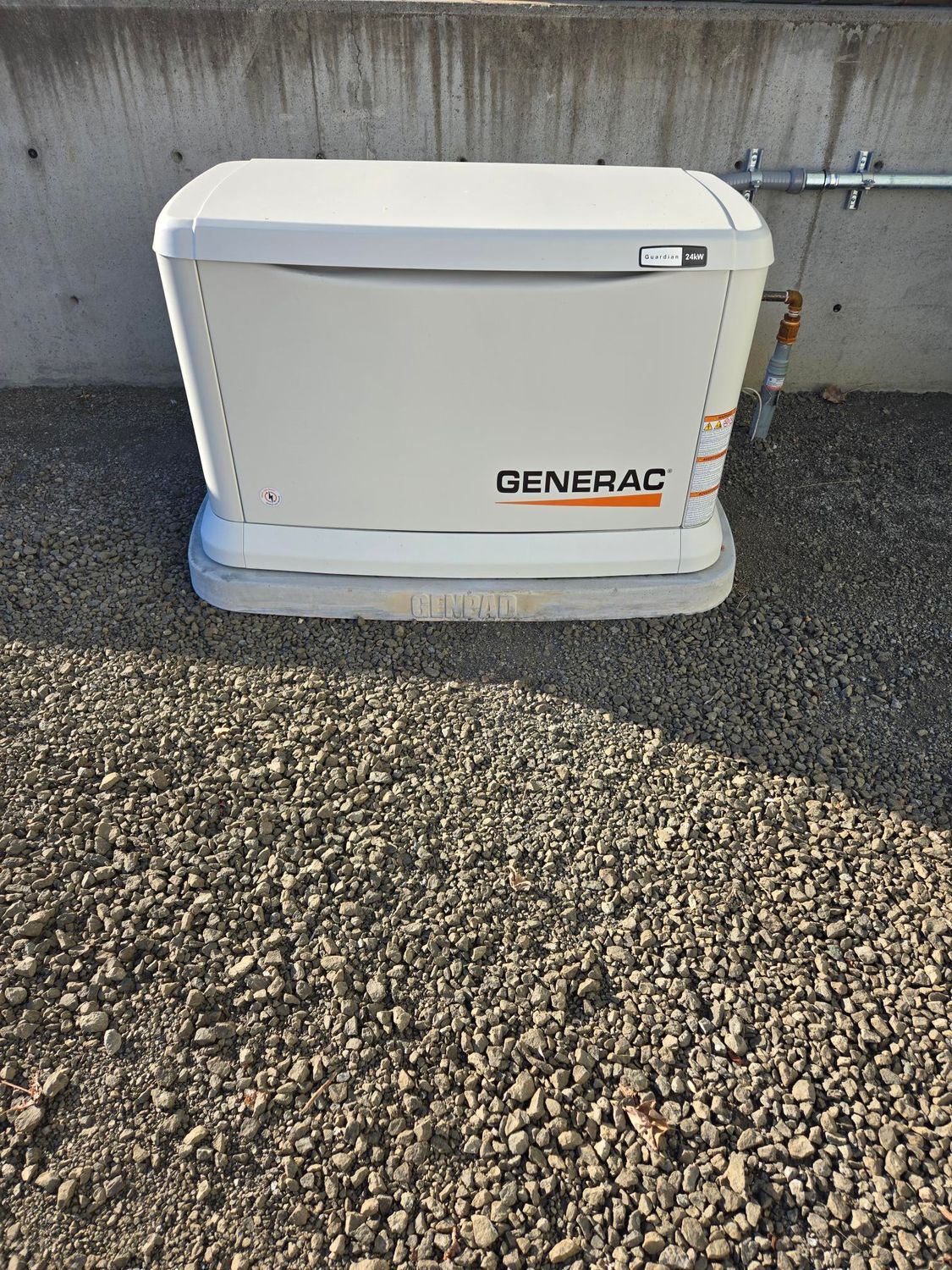 White Generac generator unit on a gravel surface beside a wall.