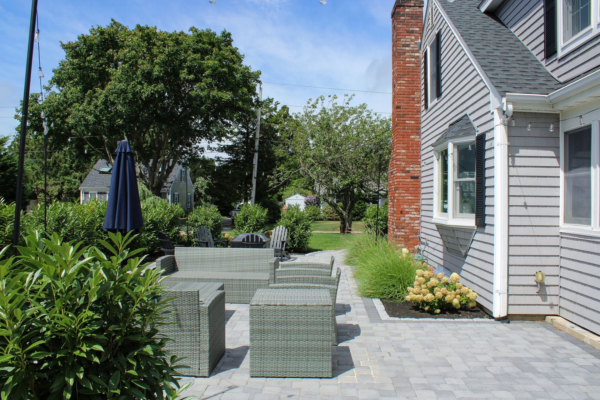 Patio with wicker furniture, house with gray siding and brick chimney, surrounded by greenery.