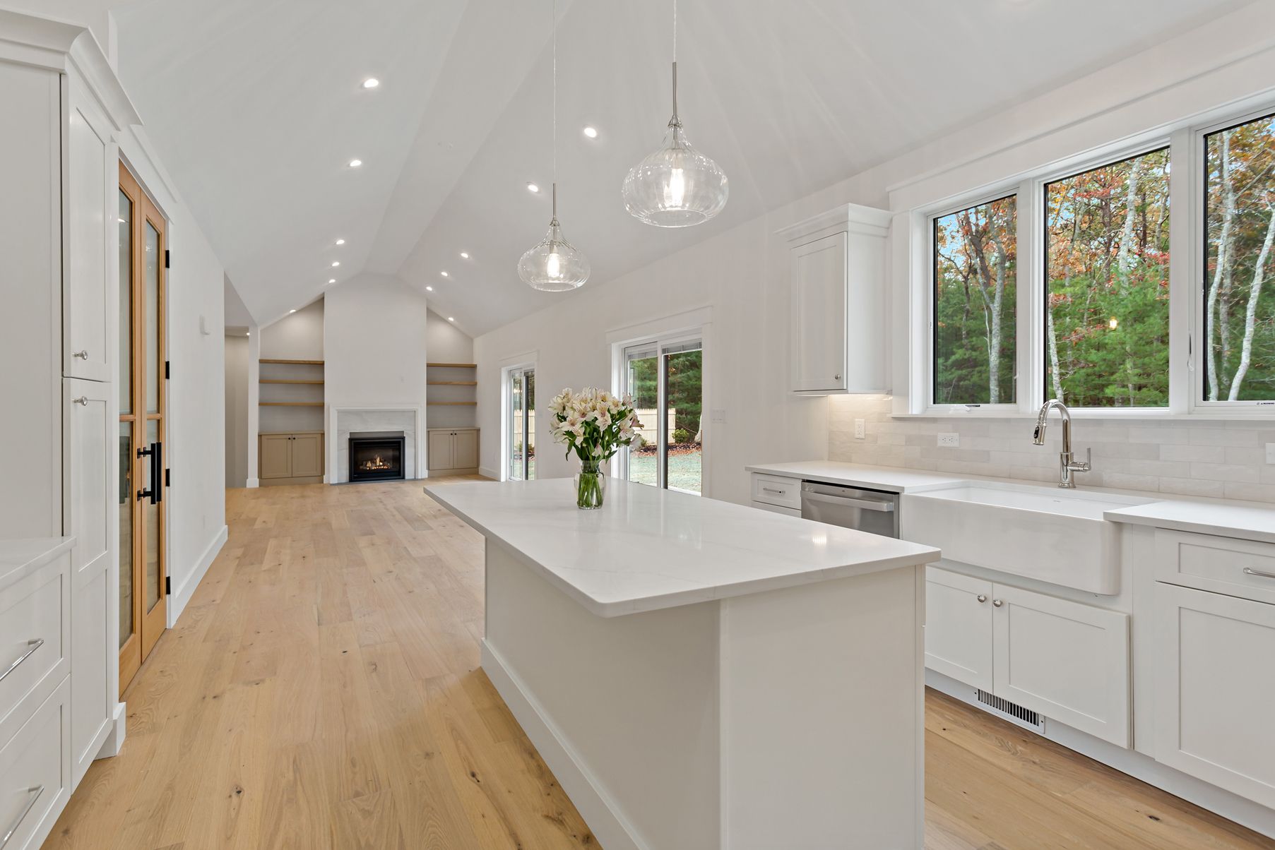 White kitchen with island, wooden floors, and large windows overlooking trees.