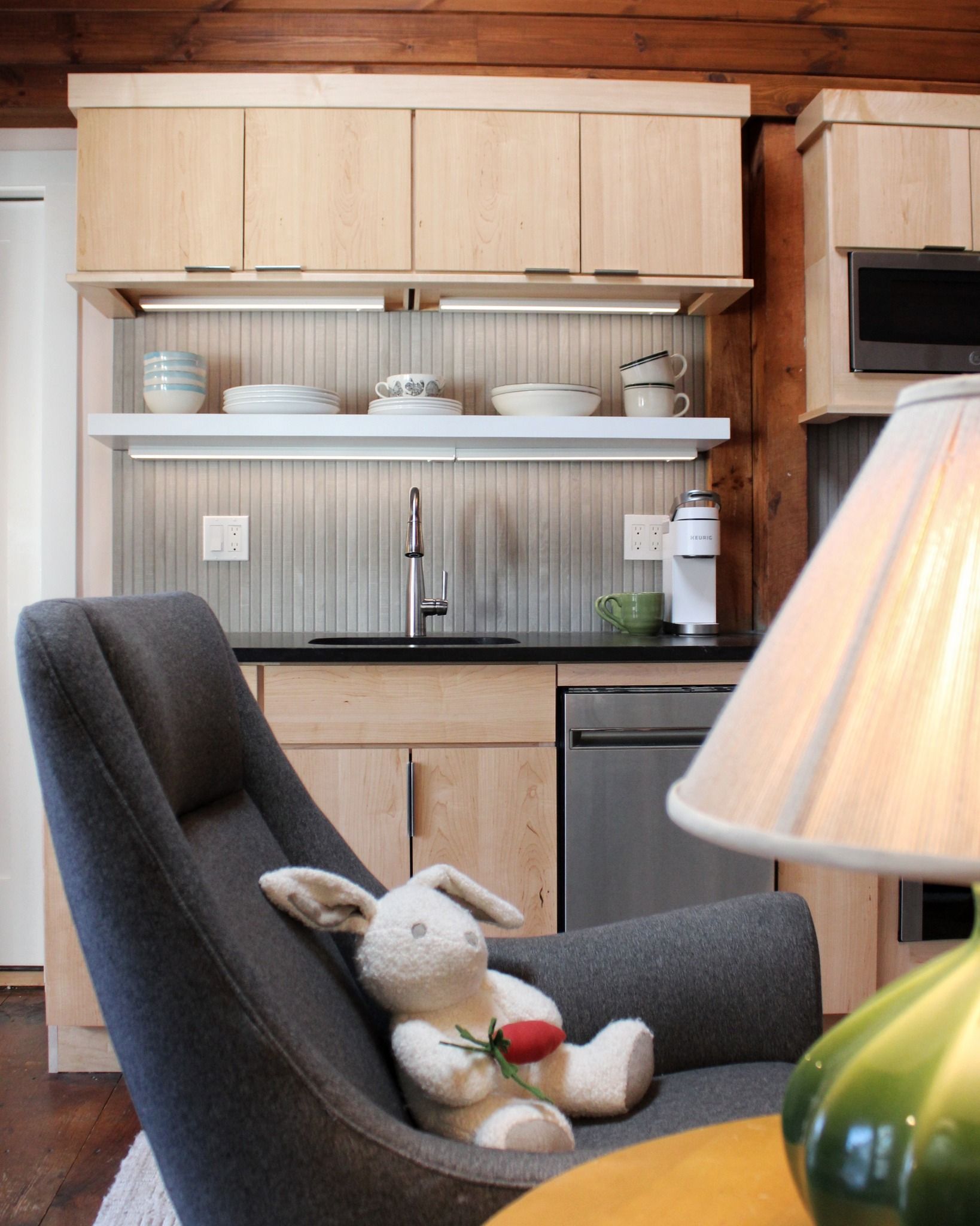 Small kitchen with light wood cabinets, black countertop, and a gray armchair with a stuffed bunny.