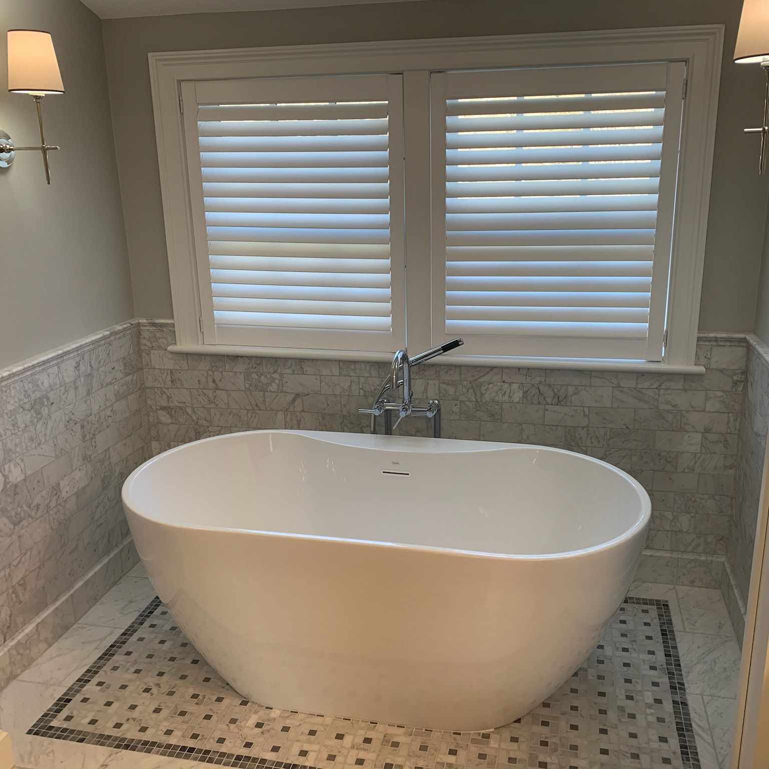 Bathroom with white oval tub, windows with blinds, gray and white tile, sconces.