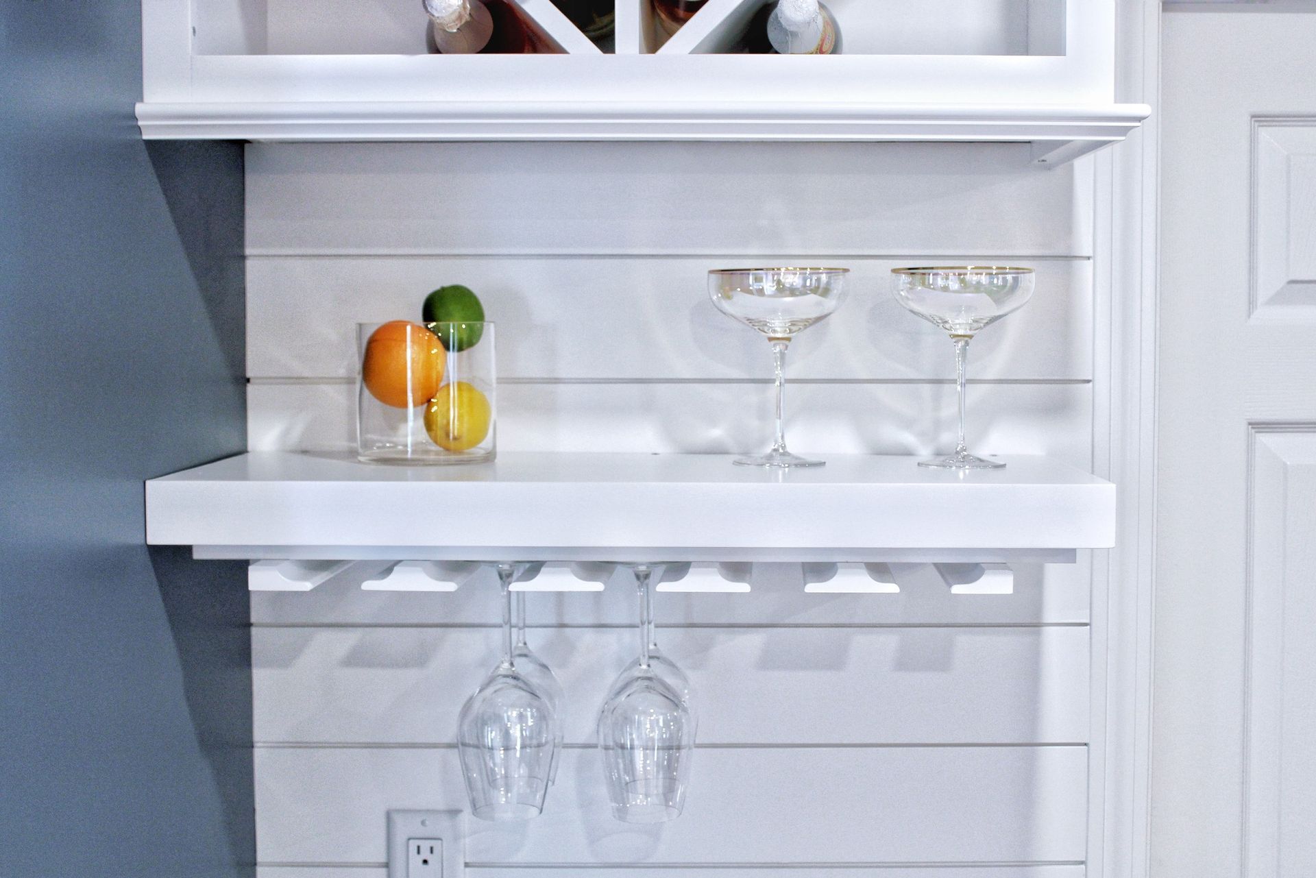 White bar shelves with wine glasses, fruit, and wine bottles.