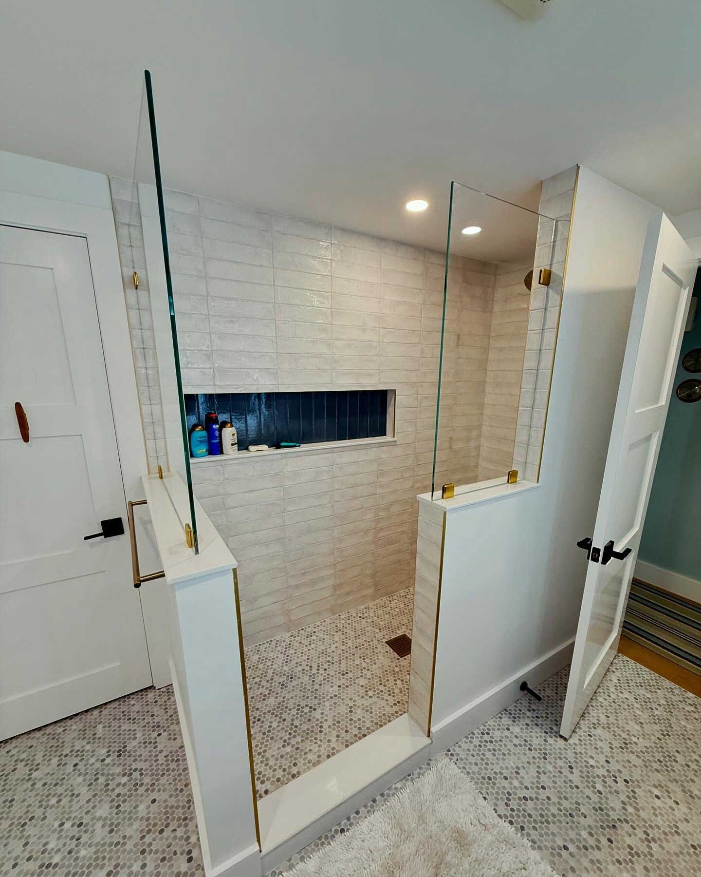 Bathroom with walk-in shower. White tiled walls, glass doors, pebble floor, built-in shelf, white cabinets, and a white door.