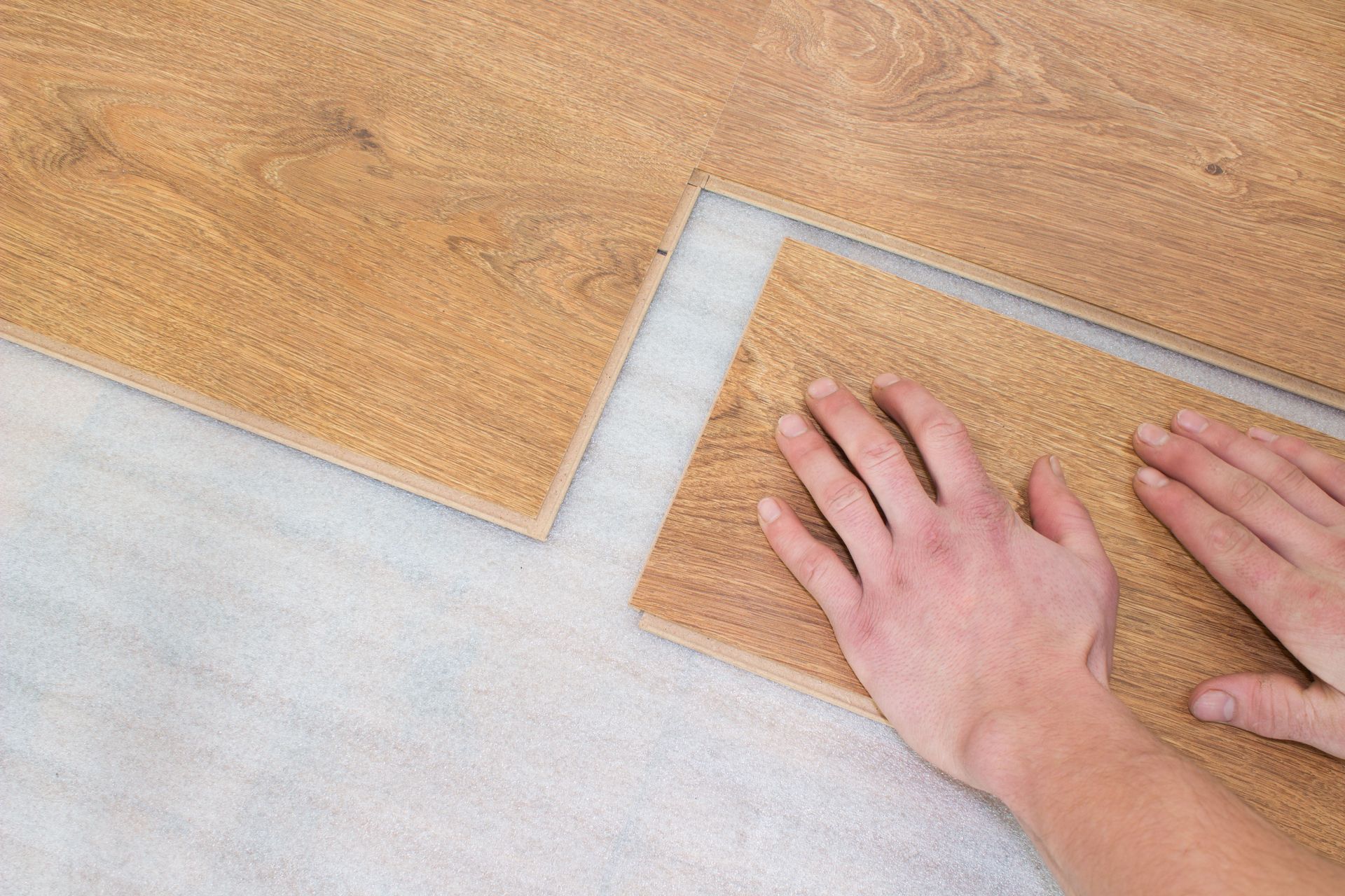 Hands installing wood-look flooring over a light-colored underlayment.