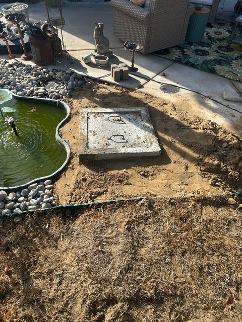 A manhole cover is sitting in the dirt next to a fountain.