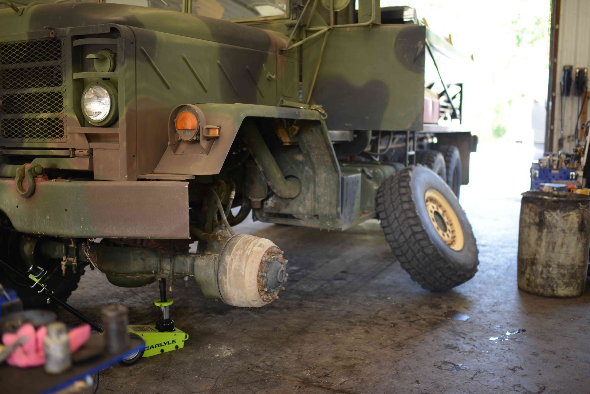 Military truck with a raised front wheel, parked inside a garage with tools on the floor.