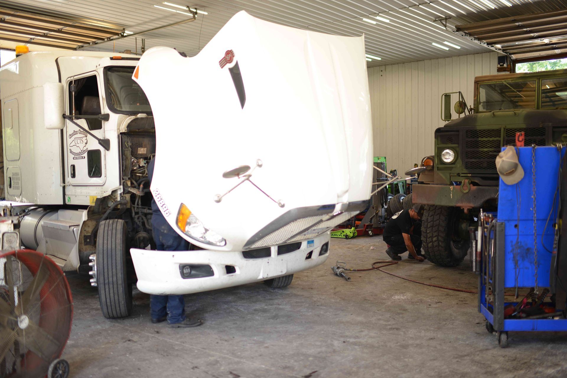 White semi-truck in a repair shop with its hood open, beside a green truck and blue equipment.